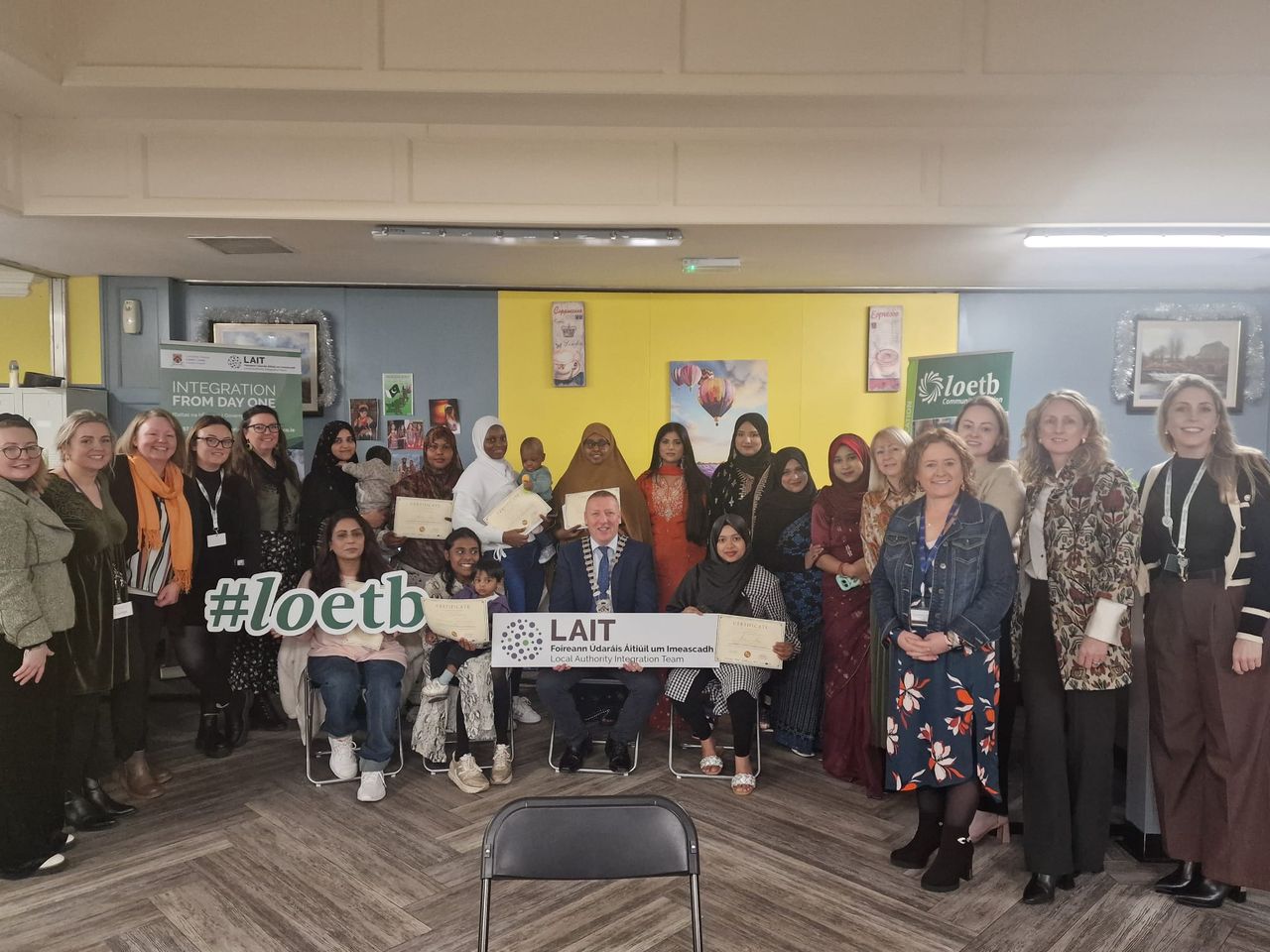 A diverse group of people holds certificates and signs, smiling in a room with colorful walls and banners for an integration event.